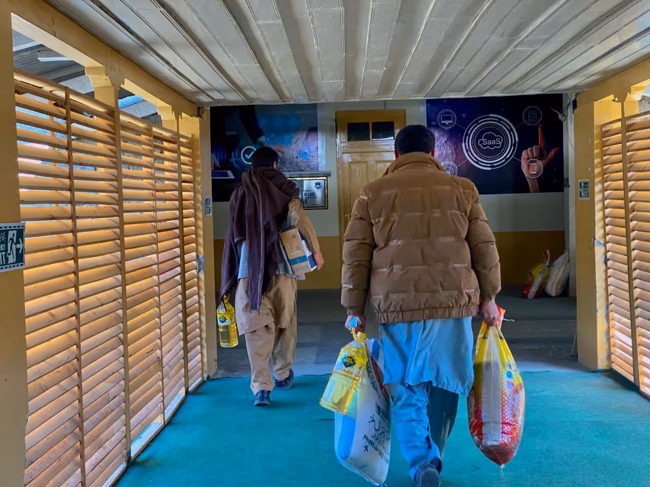 Two men walking indoors carrying grocery bags and cooking oil bottles under covered corridor.