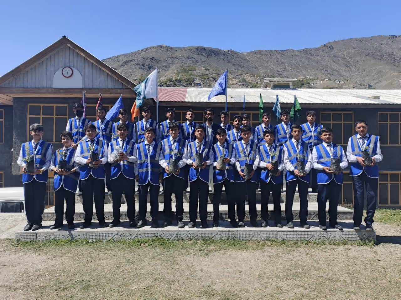 Group of young boys in matching blue vests and white shirts holding small potted plants, standing in front of a building with mountains in the background.