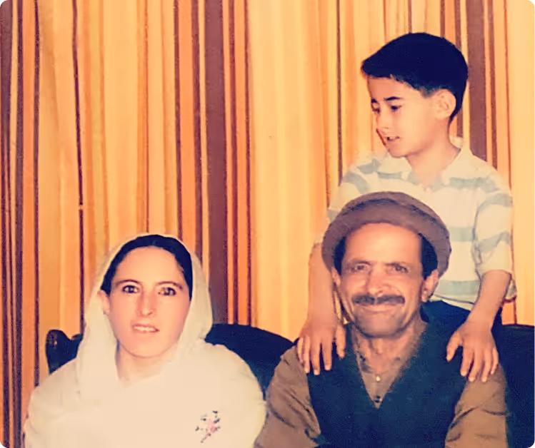 A family portrait with a woman in a white headscarf, a smiling man wearing a traditional hat, and a young boy standing behind them with his hands on the man's shoulders.