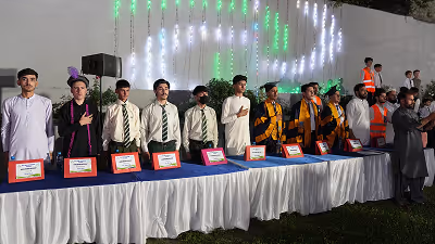 Group of young men standing behind a long table with awards, some wearing formal clothing and some in graduation gowns, at an outdoor event with decorative lights in the background.