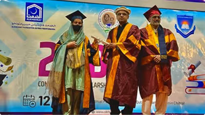 Three graduates in maroon and gold gowns and caps standing on stage during a graduation ceremony with a colorful backdrop.