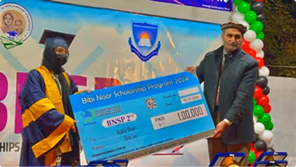 A graduate in a cap and gown receiving a large ceremonial check for the Bibi Noor Scholarship Program 2024 from a man at an event decorated with balloons.