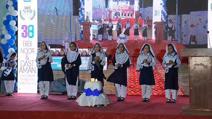 Six girls in school uniforms and headscarves standing on a stage with microphones, performing at an event with a backdrop showing more performers.
