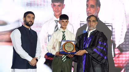 A young student in a school uniform receiving an award plaque from a man in academic regalia, with another man standing beside them on stage.