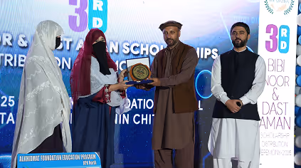 A man in traditional attire hands an award plaque to a woman wearing a maroon hijab on stage, while two other individuals stand nearby.