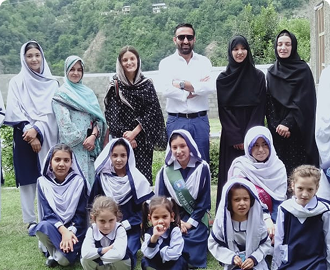 Group portrait of a man standing with a group of girls in school uniforms and headscarves, outdoors with green hills in the background.