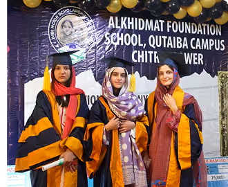 Three young women in graduation gowns and caps standing in front of a banner for Alkhidmat Foundation School, Qutaiba Campus in Chitral Tower.