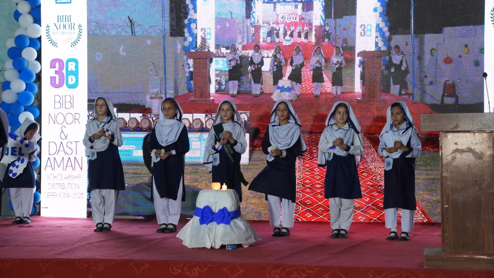 Seven schoolgirls in navy and white uniforms standing on a red stage holding candles during a school event with a decorated podium and balloon arch in the background.