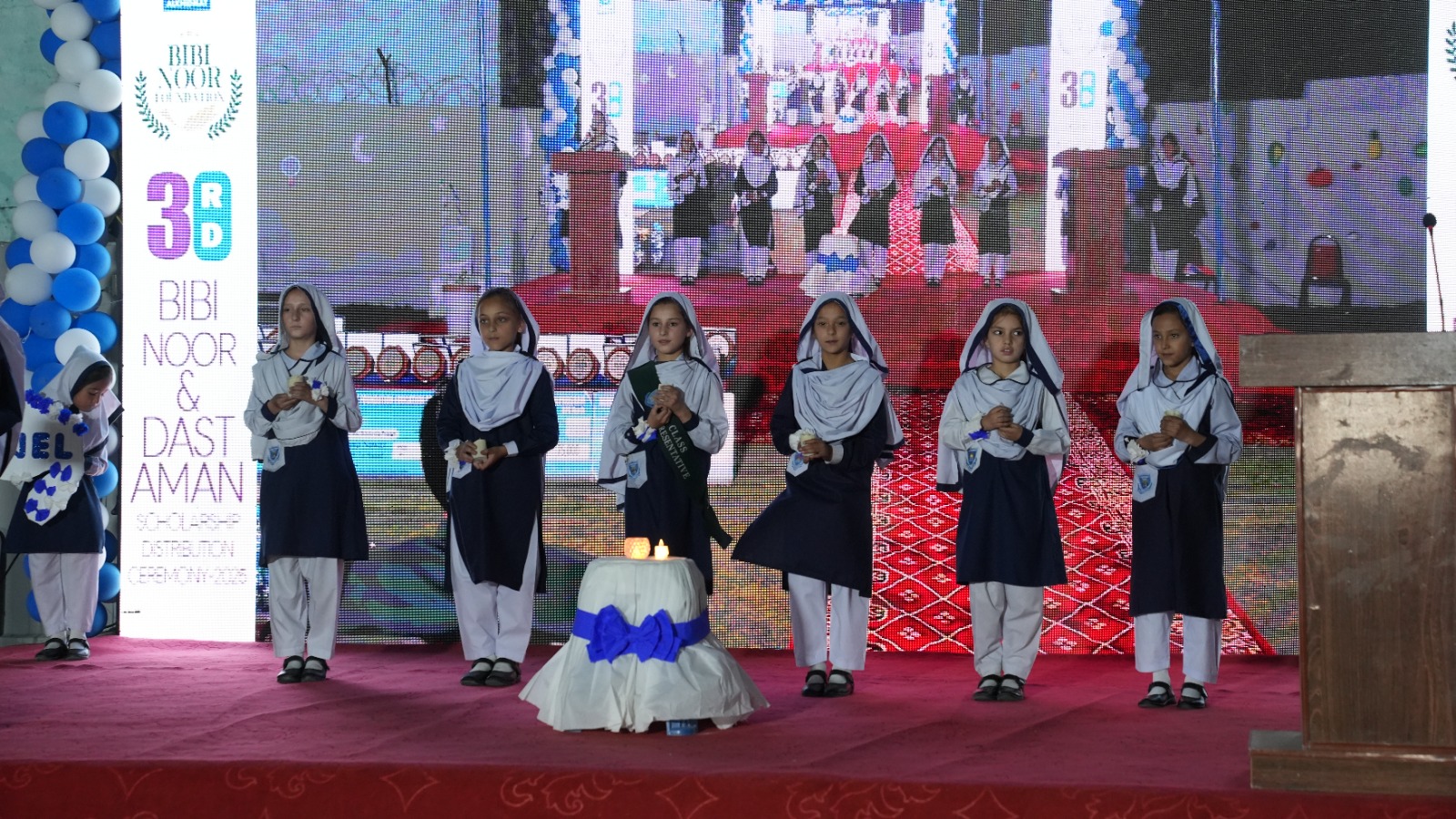 Seven schoolgirls in navy and white uniforms standing on a red stage holding candles during a school event with a decorated podium and balloon arch in the background.