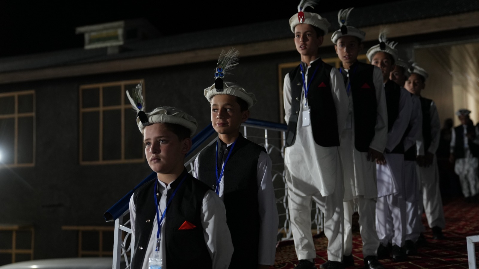 Young boys wearing traditional white outfits with black vests and feathered hats standing in a line indoors at night.