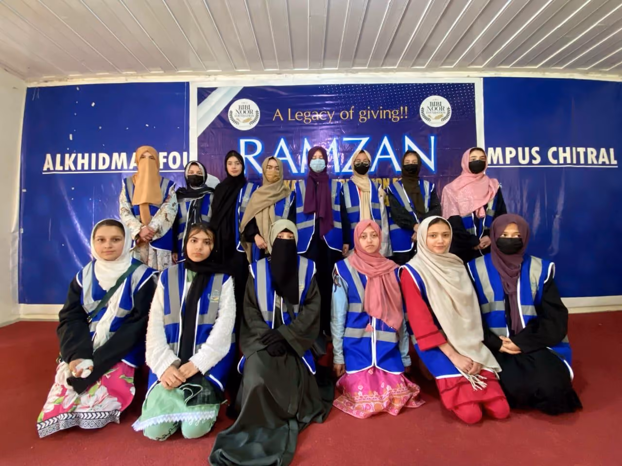 Group of young women wearing headscarves and blue reflective vests posing indoors against a backdrop with text about Ramzan and Alkhidmat Foundation.