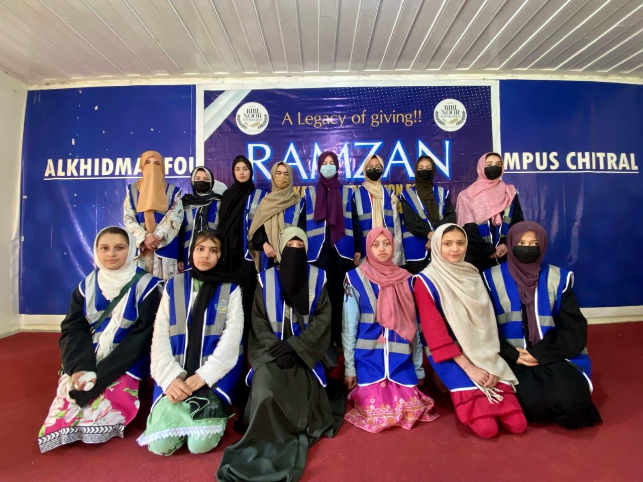 Group of young women wearing headscarves and blue reflective vests posing indoors against a backdrop with text about Ramzan and Alkhidmat Foundation.