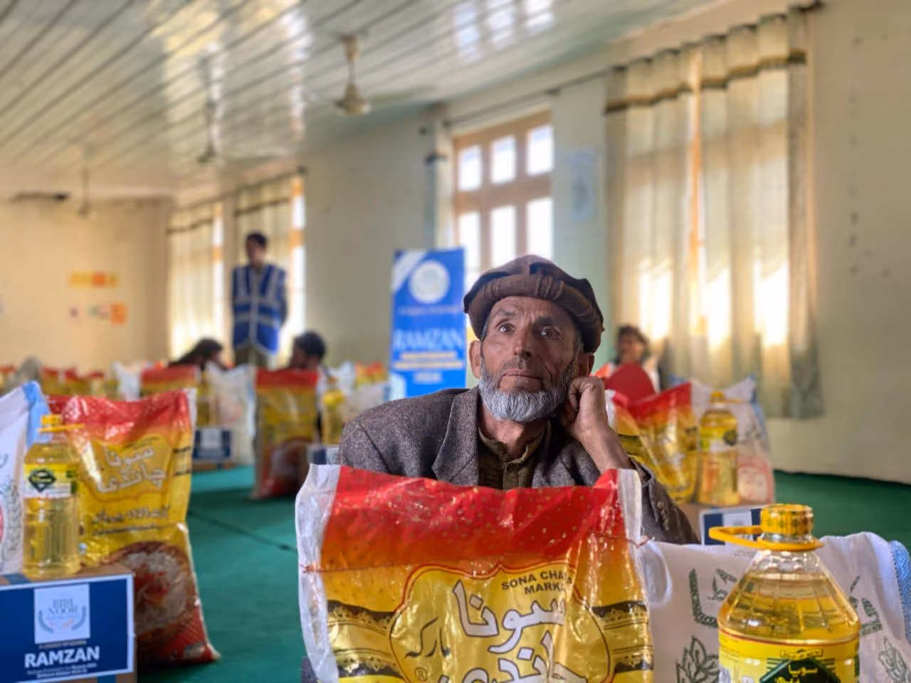 Elderly man with a beard wearing a traditional hat sitting among bags of food supplies and cooking oil in a community distribution room.
