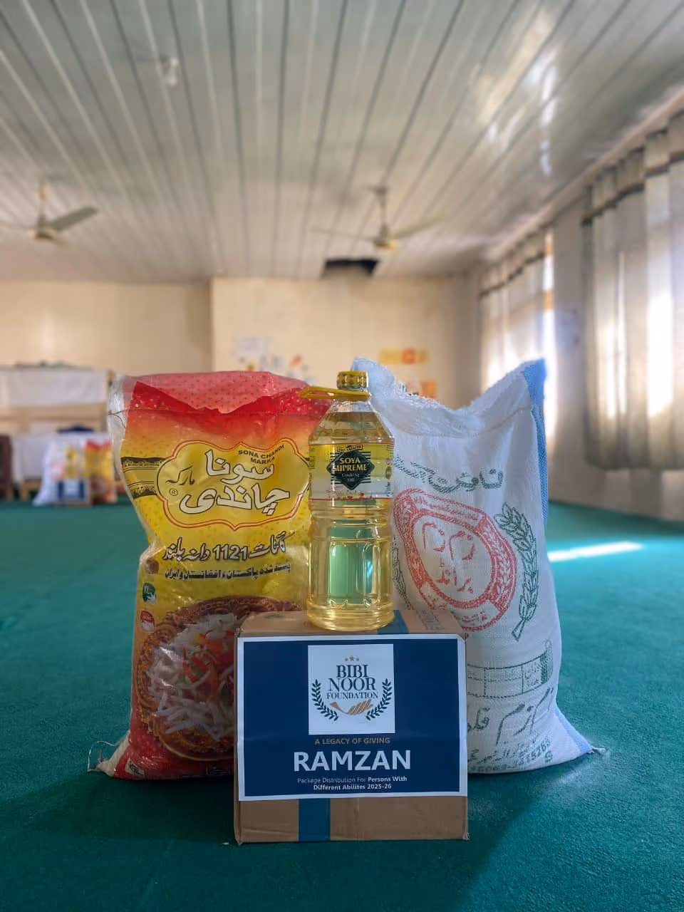 Package including rice, cooking oil, and a box labeled 'Bibi Noor Foundation Ramzan Package Distribution for Persons with Different Abilities 2025-26' on a green carpeted floor inside a room.