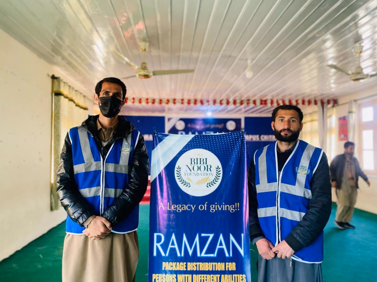 Two men wearing blue reflective vests standing beside a banner for Bibi Noor Foundation's Ramzan package distribution for persons with different abilities.