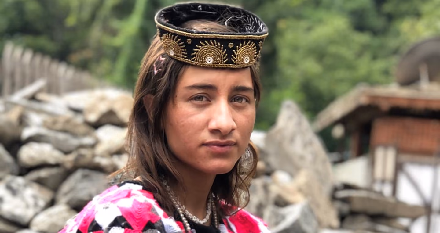 Portrait of a woman wearing traditional attire with a decorative black and gold headpiece and a pink patterned shawl, with a rocky background.