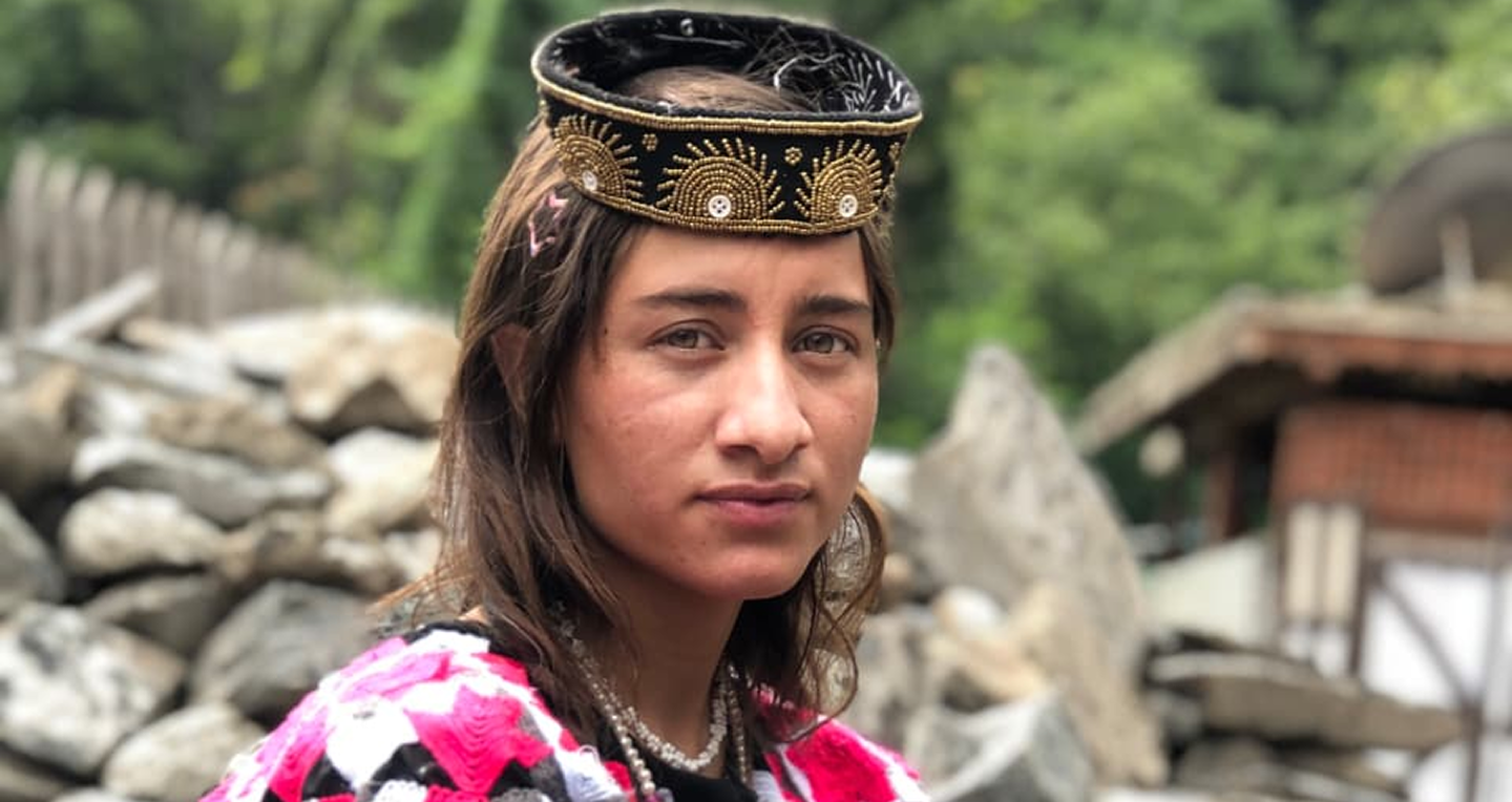 Portrait of a woman wearing traditional attire with a decorative black and gold headpiece and a pink patterned shawl, with a rocky background.