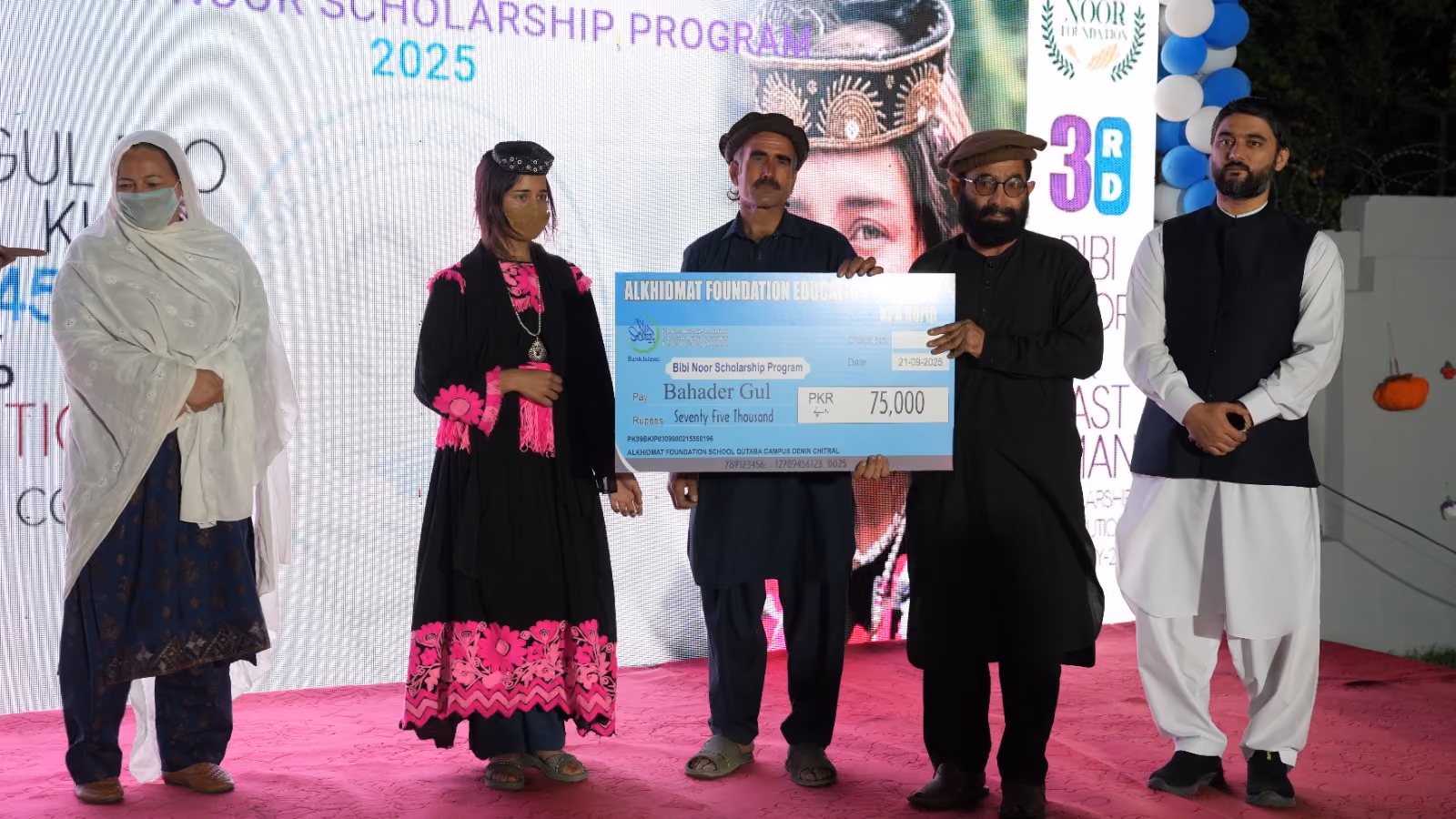 Five people standing on a stage, with one man holding a large blue check for 75,000 PKR from the Bibi Noor Scholarship Program.