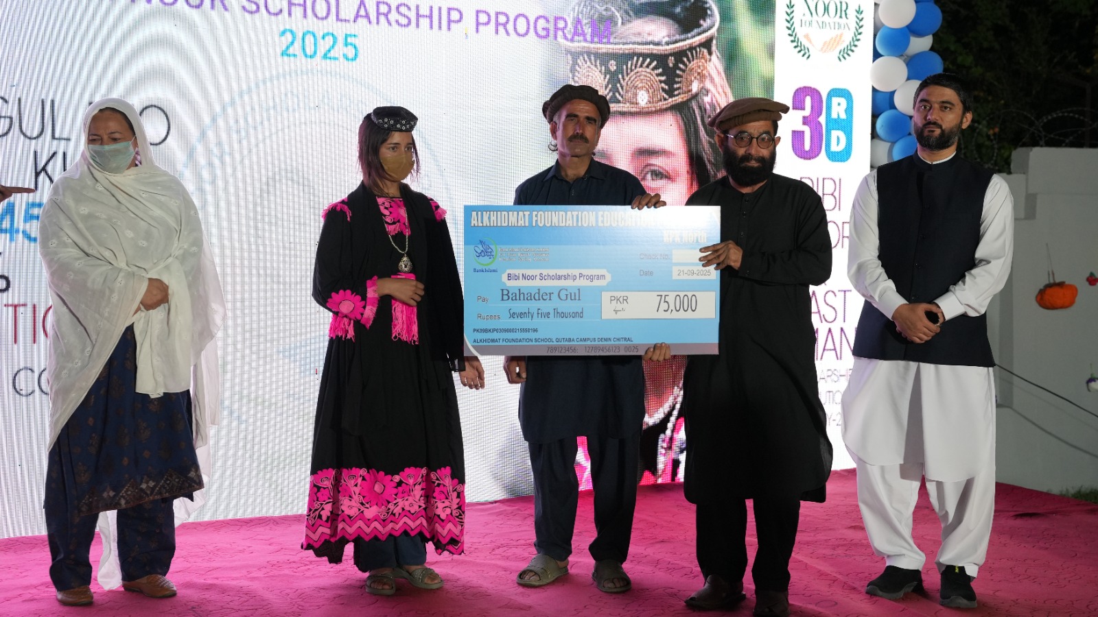 Five people standing on a stage, with one man holding a large blue check for 75,000 PKR from the Bibi Noor Scholarship Program.