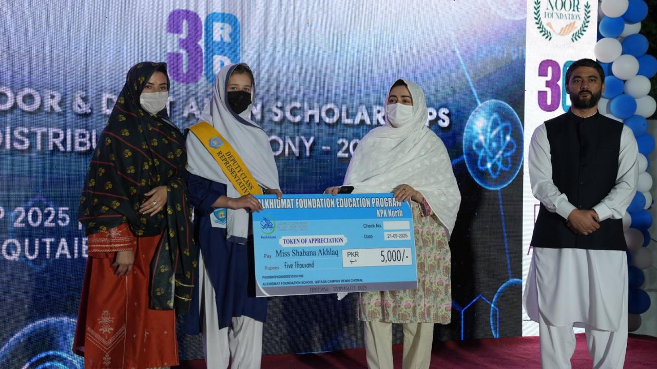 Four people stand on a stage during a scholarship award ceremony, with one woman holding a large check for 5,000 PKR presented to Miss Shabana Akhlaq.
