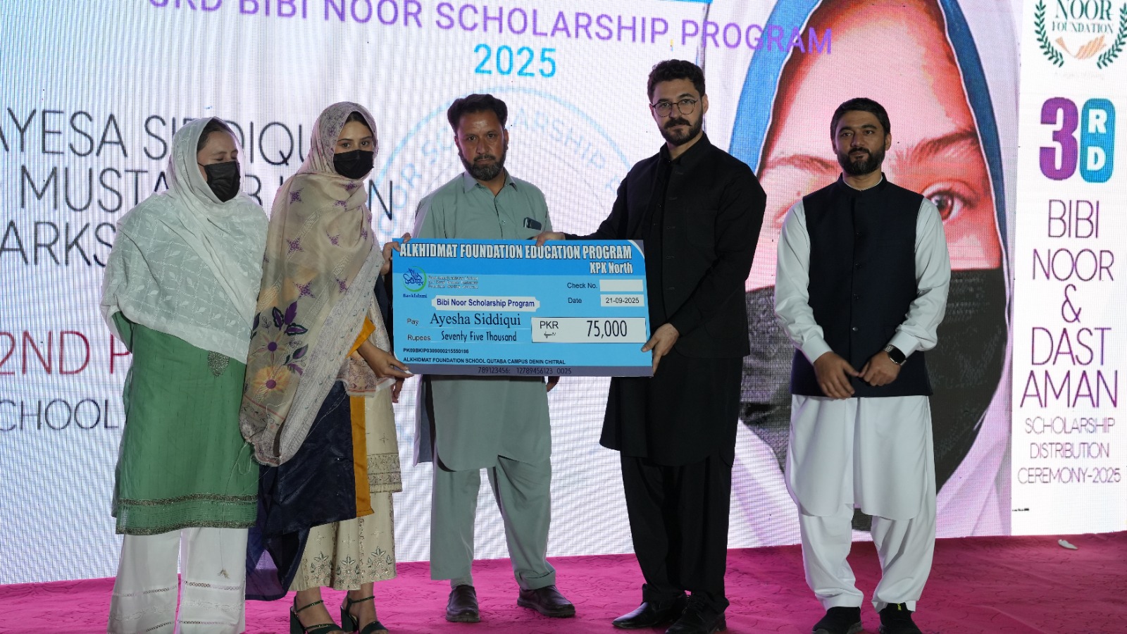 Five people standing on stage during the 3rd Bibi Noor & Dast Aman Scholarship Distribution Ceremony 2025, holding a large check for 75,000 PKR awarded to Ayesha Siddiqui.
