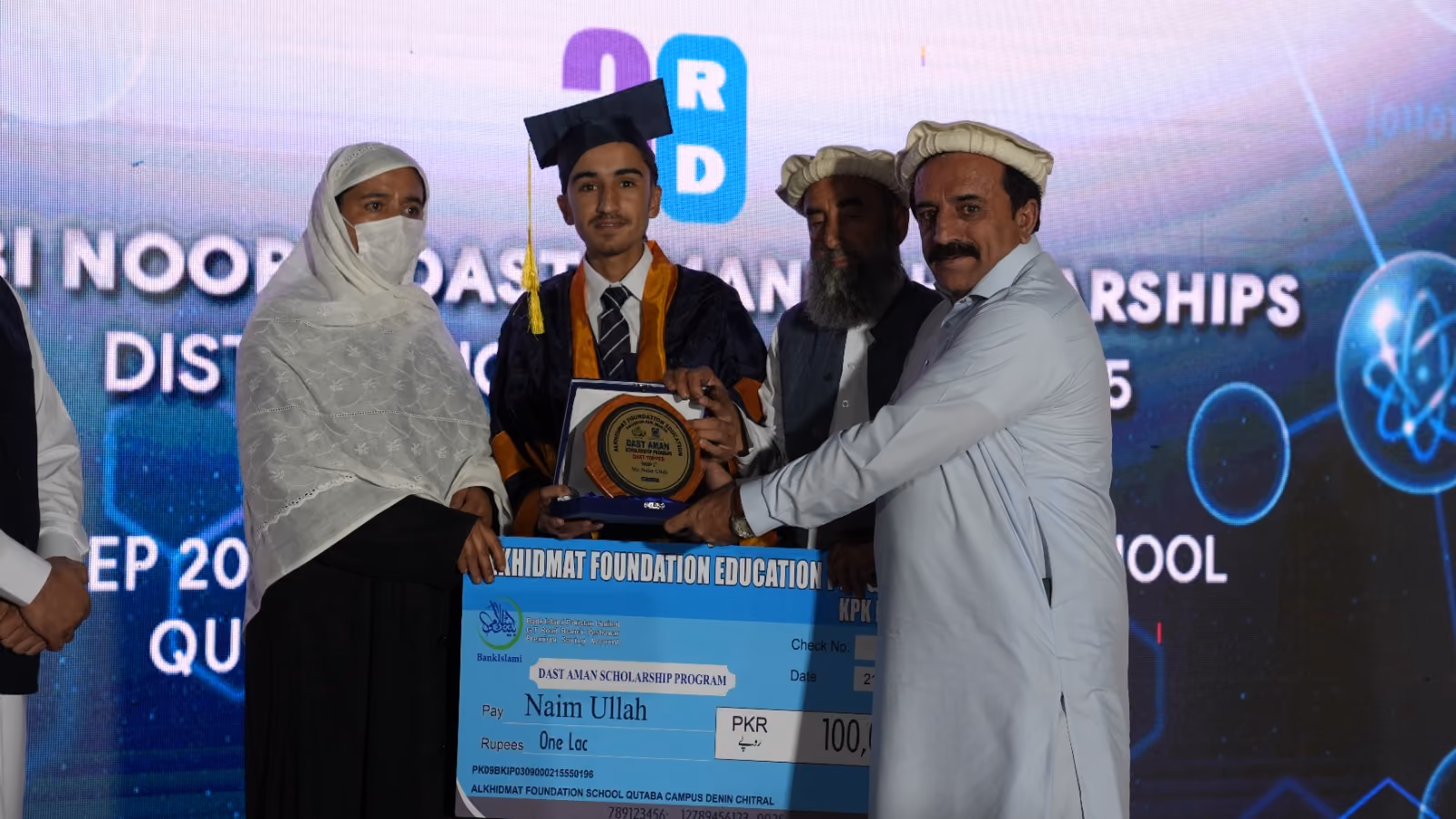 Young man in graduation cap and gown holding a scholarship plaque and oversized check worth 100,000 PKR with three adults around him on stage.