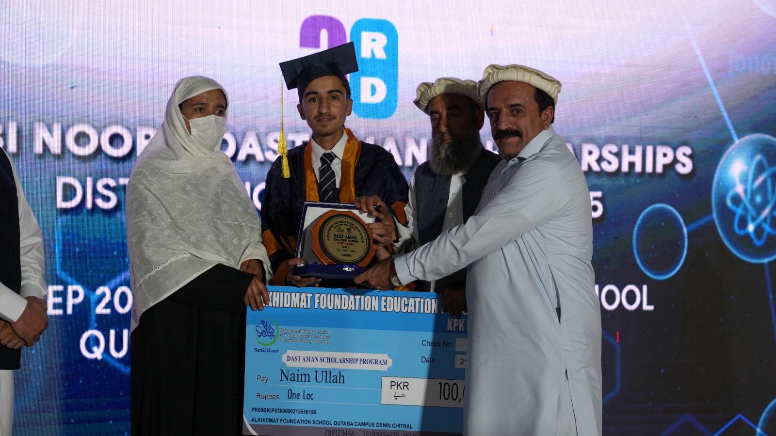 Young man in graduation cap and gown holding a scholarship plaque and oversized check worth 100,000 PKR with three adults around him on stage.