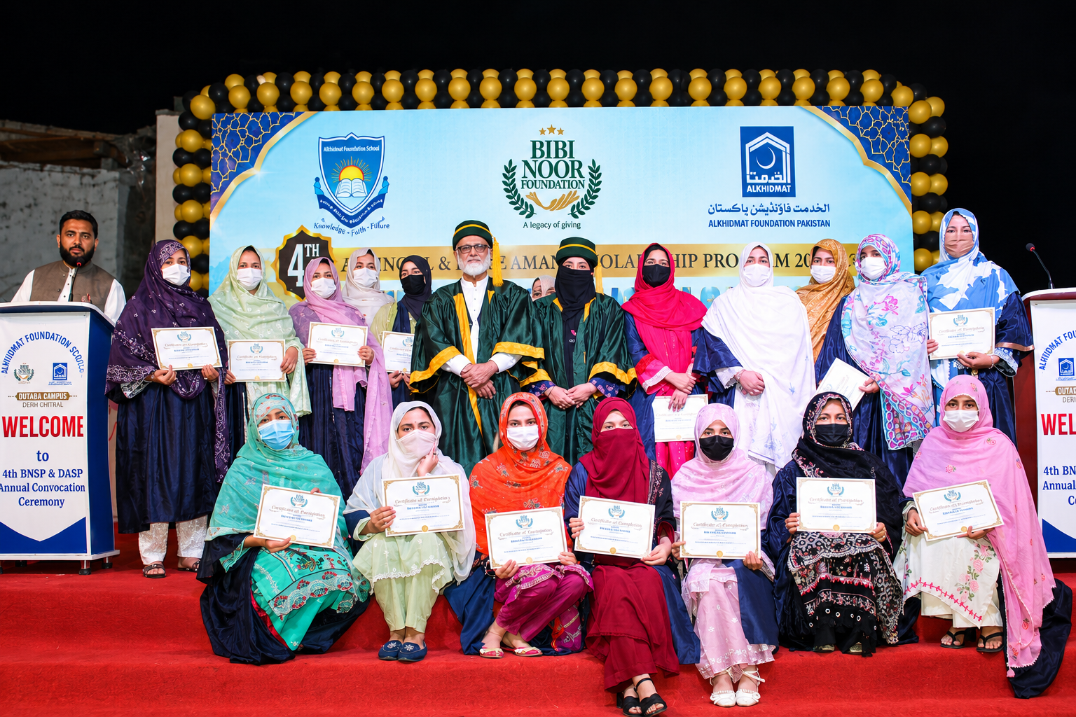 Group portrait of men and women at an annual convocation ceremony, with participants holding certificates; two individuals in the center wear green academic gowns and caps. The background features logos of Bibi Noor Foundation and Alkhidmat Foundation Pakistan, along with text about the 4th BNSP & DASP convocation. The participants wear colorful cultural attire with some wearing face masks, posed on a red-carpeted stage decorated with black and yellow balloons.