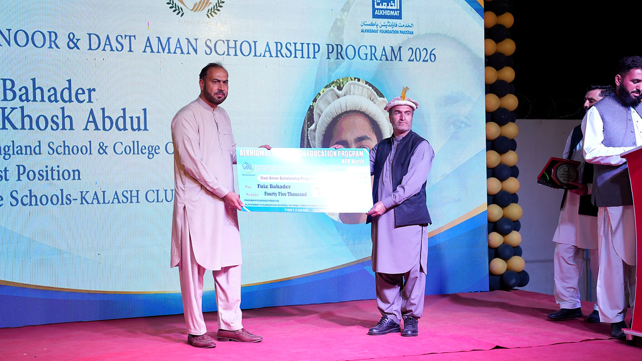 Two men standing on stage holding a large ceremonial check for Faiz Bahader worth forty-five thousand rupees during the Dast Aman Scholarship Program 2026 event with a blue and white backdrop.