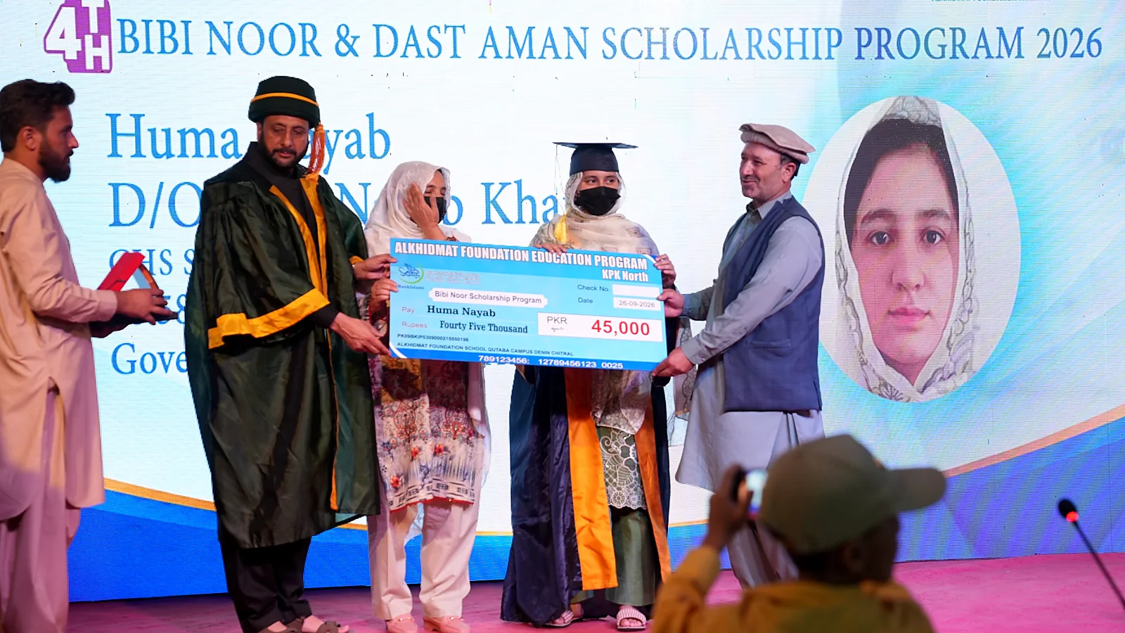 Four people on stage during a scholarship award ceremony with one woman in graduation attire holding a large ceremonial check for 45,000 PKR, awarded to Huma Nayab, with a large screen in the background displaying scholarship information and a portrait of the awardee.