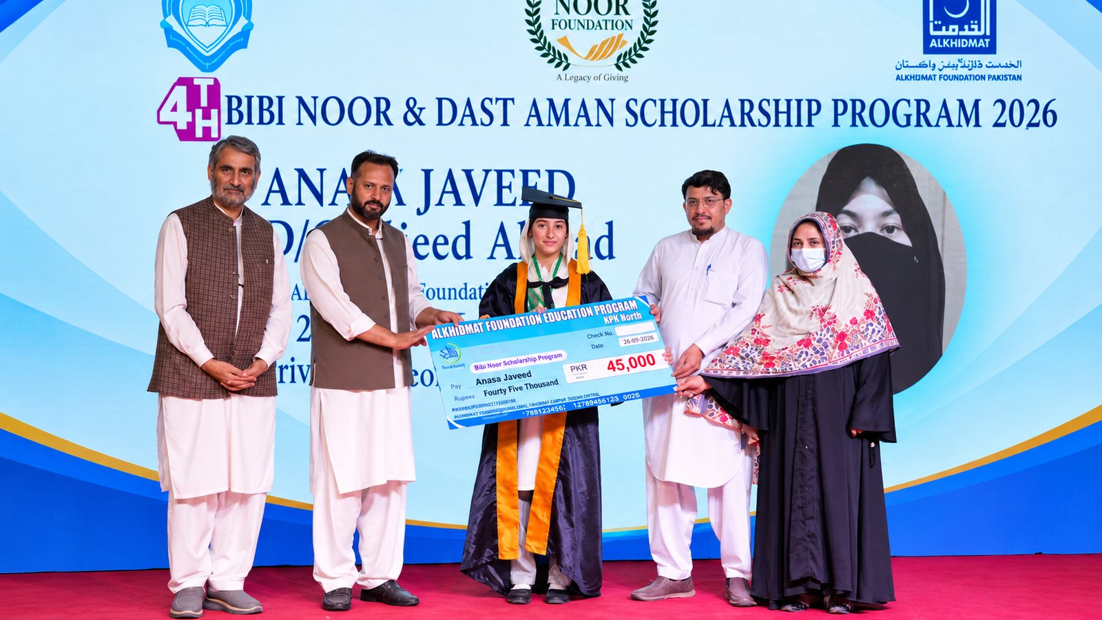 A female graduate in cap and gown holding a large ceremonial check for 45,000 PKR from the Bibi Noor & Dast Aman Scholarship Program 2026, standing with four adults on a stage with a blue backdrop displaying scholarship program text and logos.