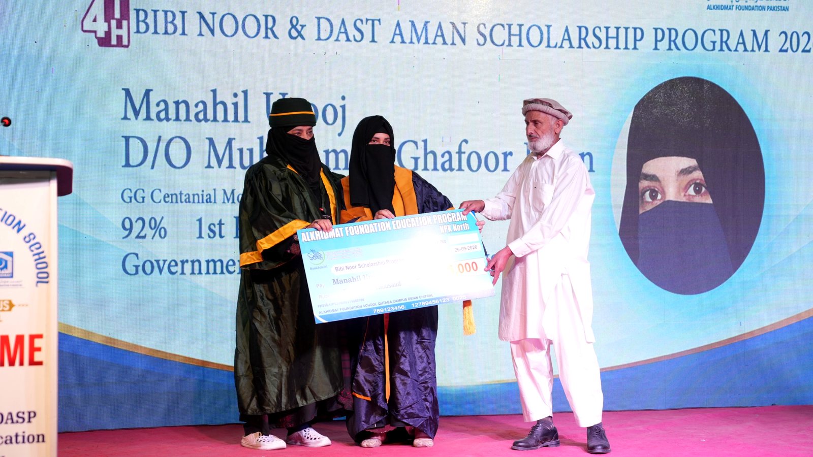 Three people on stage during a scholarship award ceremony, two women wearing black abayas and niqabs holding a large ceremonial check, and a man in white traditional attire and a cap handing it to them, against a backdrop displaying scholarship details including the name Manahil Usooj and academic achievements.