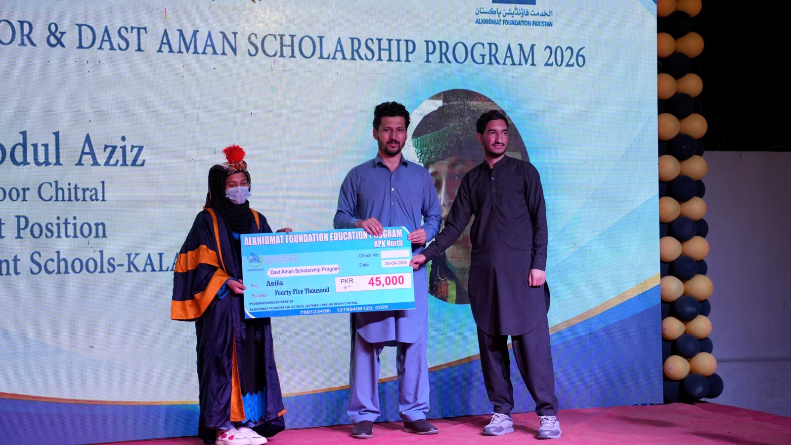 Three people standing on a stage during a scholarship award ceremony; a young woman in a graduation gown and face mask holds a large blue check for 45,000 PKR from Alkhidmat Foundation Education Program, flanked by two men in traditional Pakistani attire, with a backdrop displaying scholarship program details and decorative balloon columns on the right.