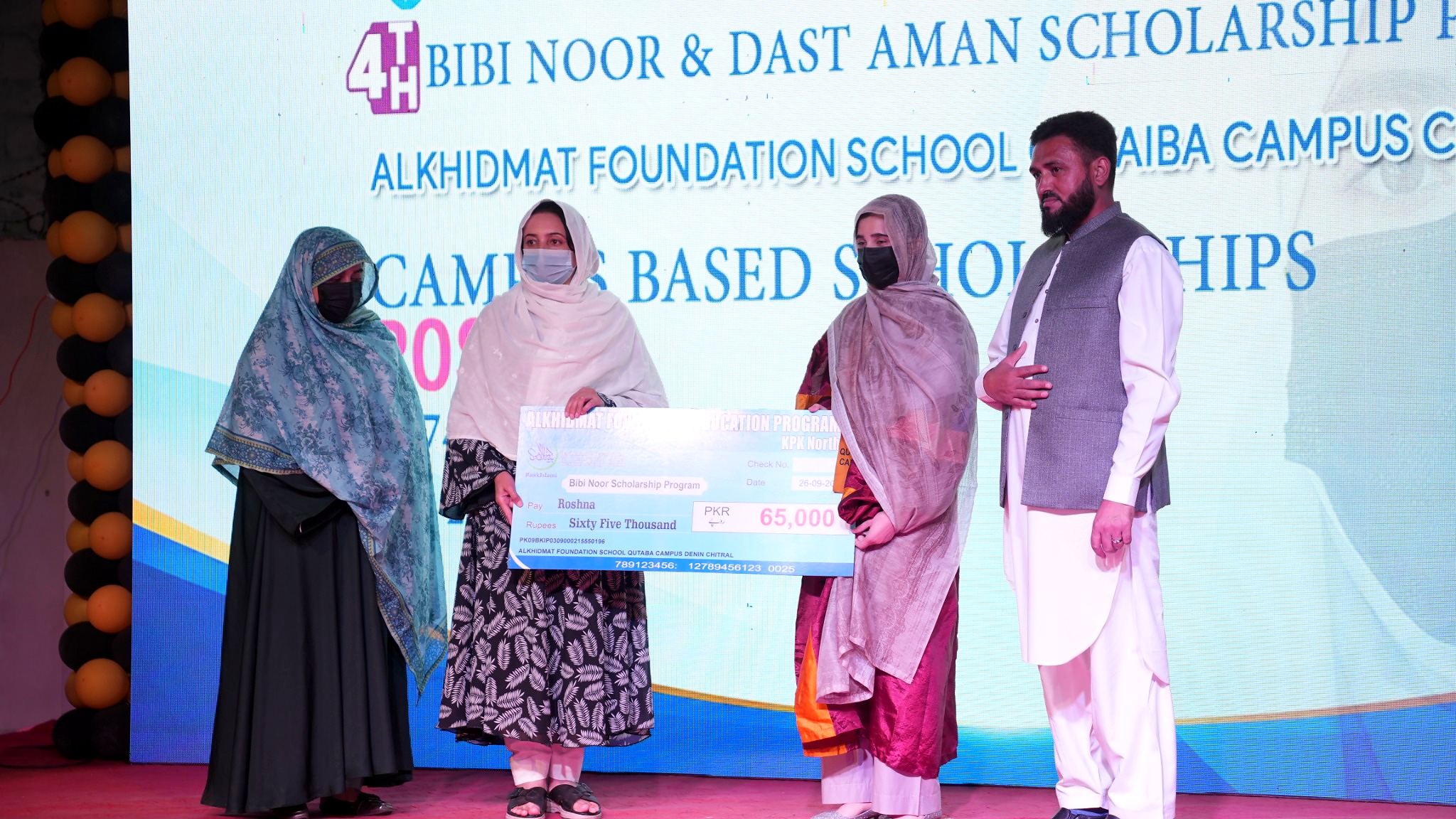 Four people standing on a stage during an award ceremony, with two women wearing face masks holding a large cheque for 65,000 PKR from the Bibi Noor Scholarship Program; the others stand beside them with a backdrop displaying scholarship program information.