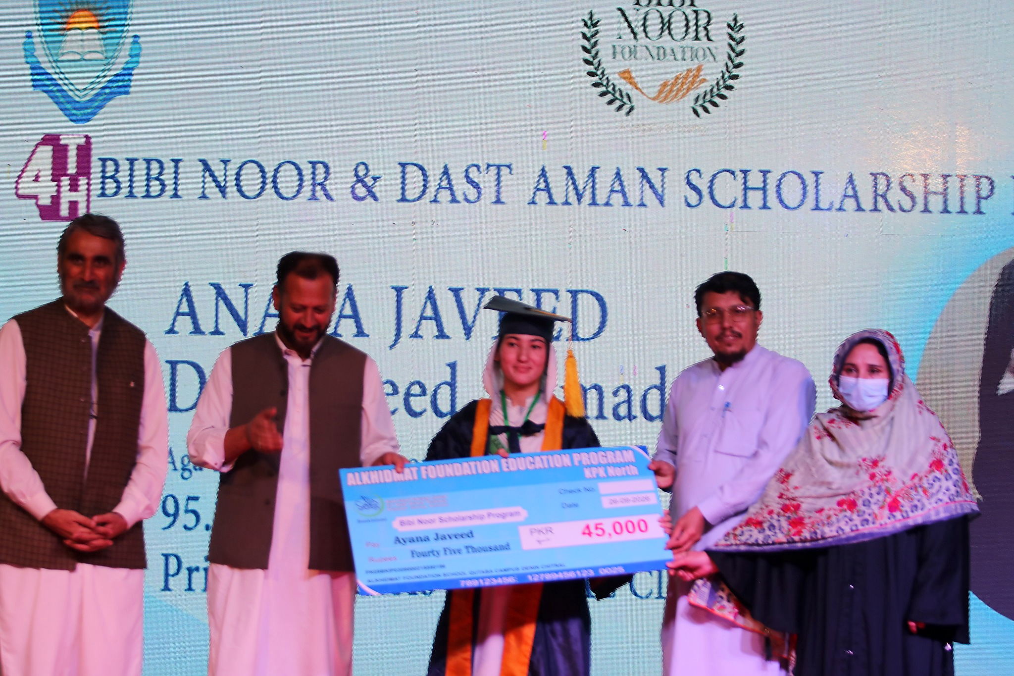 A young woman in a graduation cap and gown holds a large check for 45,000 PKR from the Alkhidmat Foundation Education Program, surrounded by four adults at a scholarship event.