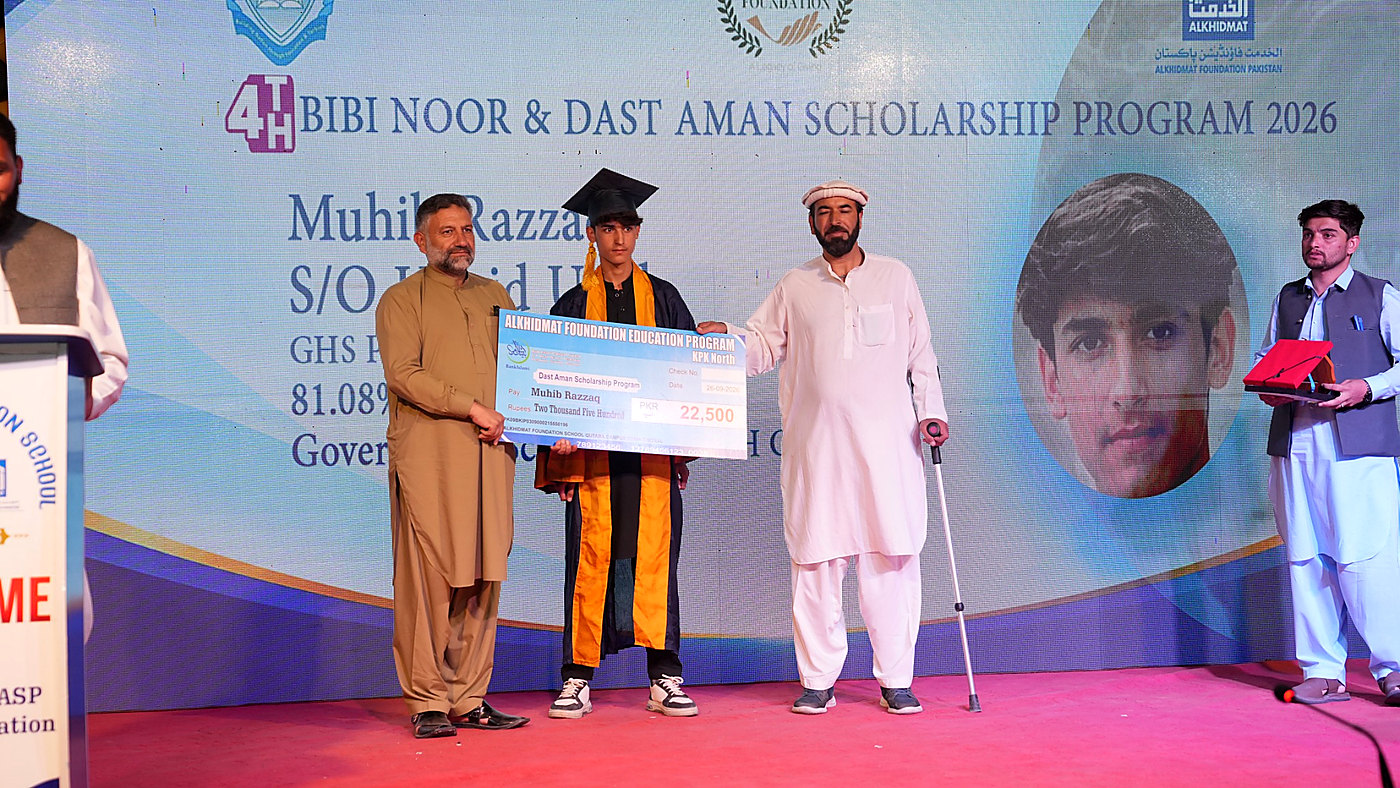 A young man in a graduation cap and gown holding a large ceremonial check for PKR 22,500 with two men standing beside him on a stage during the 4th Bibi Noor & Dast Aman Scholarship Program 2026 event.