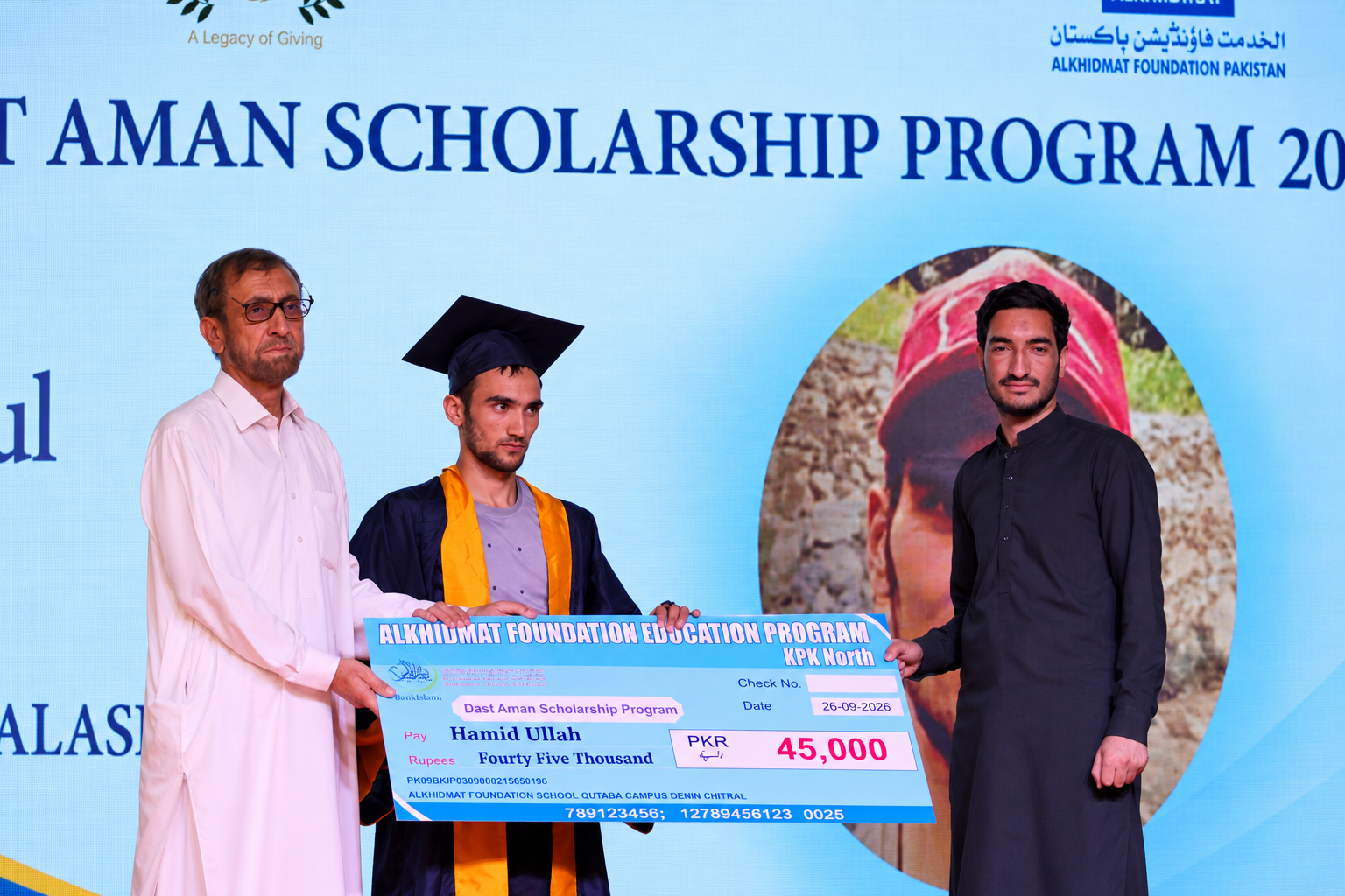 Three men on stage during the Dast Aman Scholarship Program ceremony; the man in the center wears a graduation cap and gown, holding a large ceremonial cheque for 45,000 PKR made out to Hamid Ullah, with another man on each side supporting the cheque.