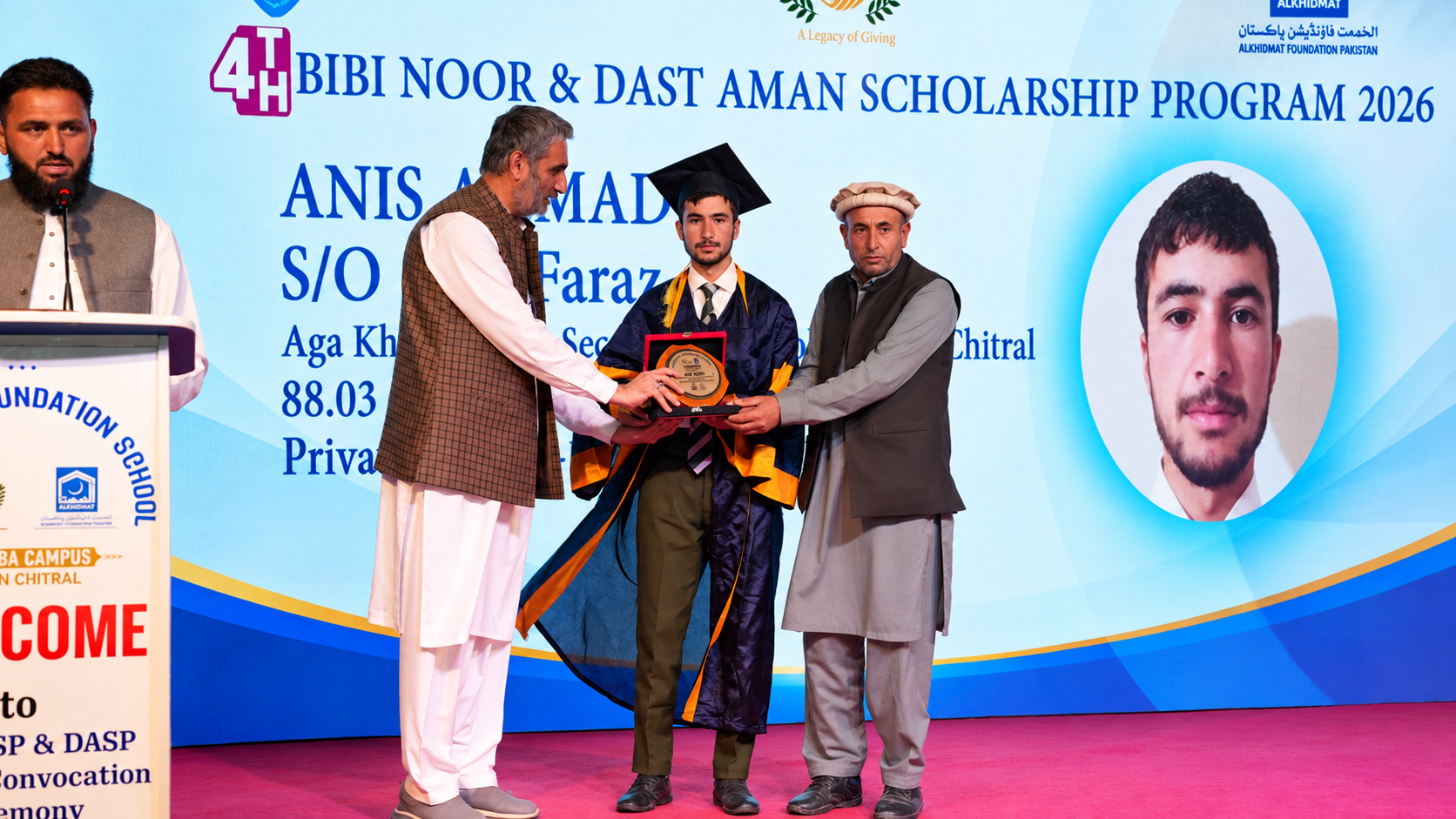 A young man in graduation gown and cap, Anis Ahmad, receiving a scholarship award plaque from two men in traditional attire on stage with a large presentation screen behind them displaying his name, photo, and scholarship details for the Bibi Noor & Dast Aman Scholarship Program 2026.