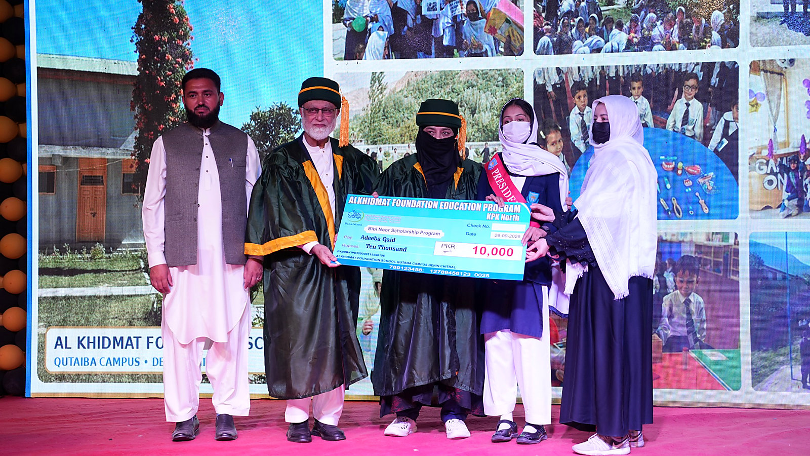Five people standing on a stage holding a large blue check for 10,000 PKR, awarded as the Bibi Noor Scholarship Program by Alkhidmat Foundation Education Program, KPK North. Two men wear academic gowns and caps, one man in traditional attire, and two women in school uniform and hijab, all wearing face masks. A large screen in the background shows images of school activities and the text 'AL KHIDMAT FOUNDATION SCHOOL QUTAIBA CAMPUS'.