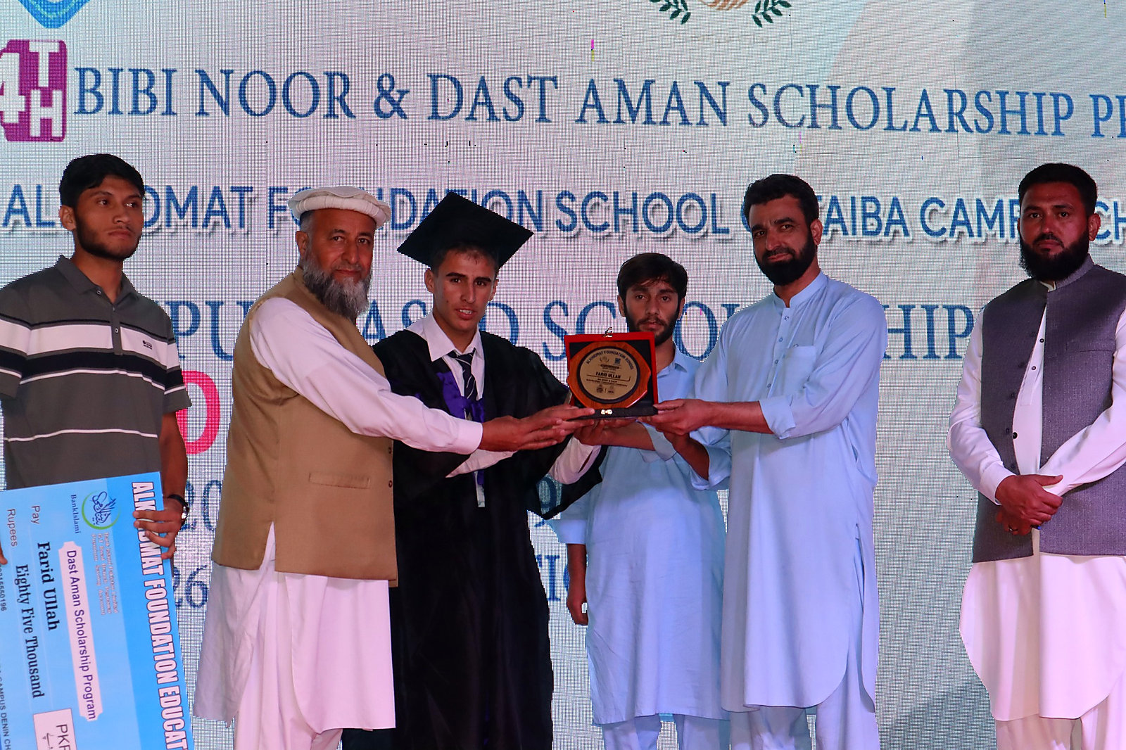Group of six men standing on stage at a scholarship event; one man in the center wearing a graduation cap and gown is holding a plaque with two others, while the man on the left holds a large blue scholarship check.