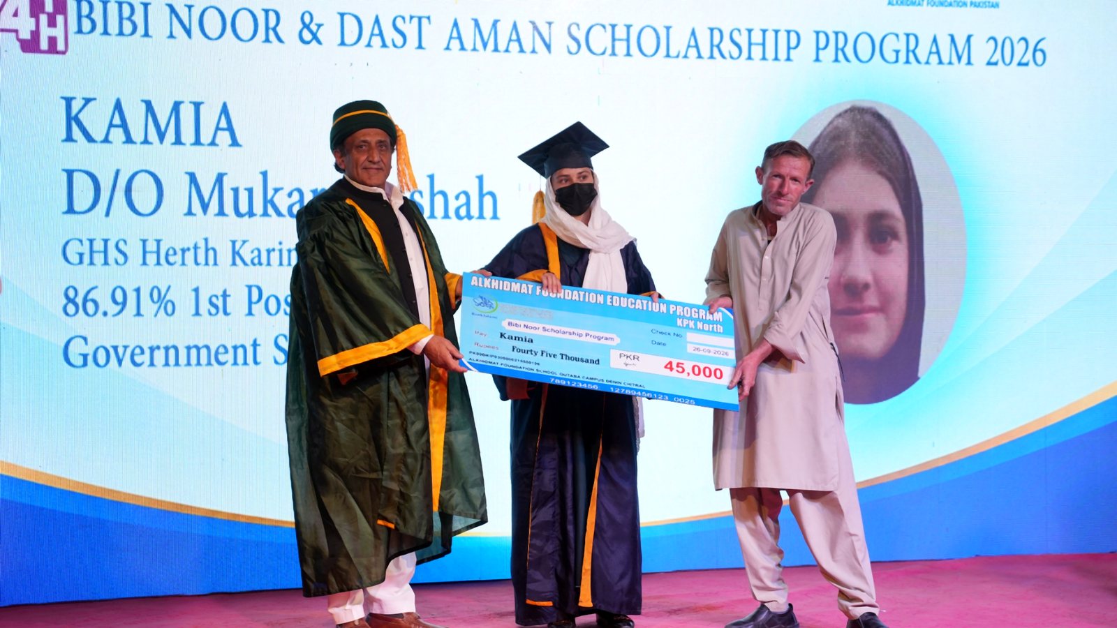 A young woman in graduation gown and cap holding a large ceremonial cheque for 45,000 PKR, standing on stage between two men; the man on the left wears academic regalia, the man on the right is in traditional attire. The backdrop shows text about the Bibi Noor & Dast Aman Scholarship Program 2026 and the woman's name Kamia, with her academic achievement of 86.91% first position.