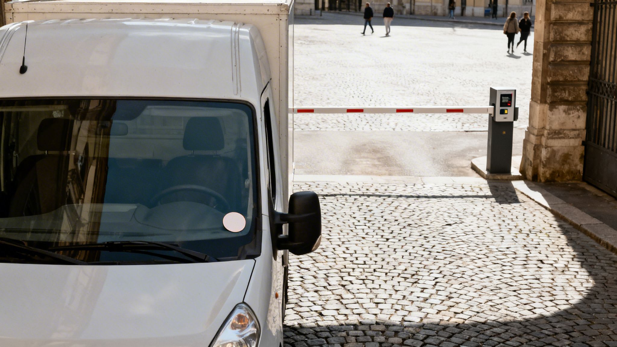 Furgone bianco fermo davanti a una barriera di accesso automatica in una piazza pavimentata, con persone sullo sfondo.