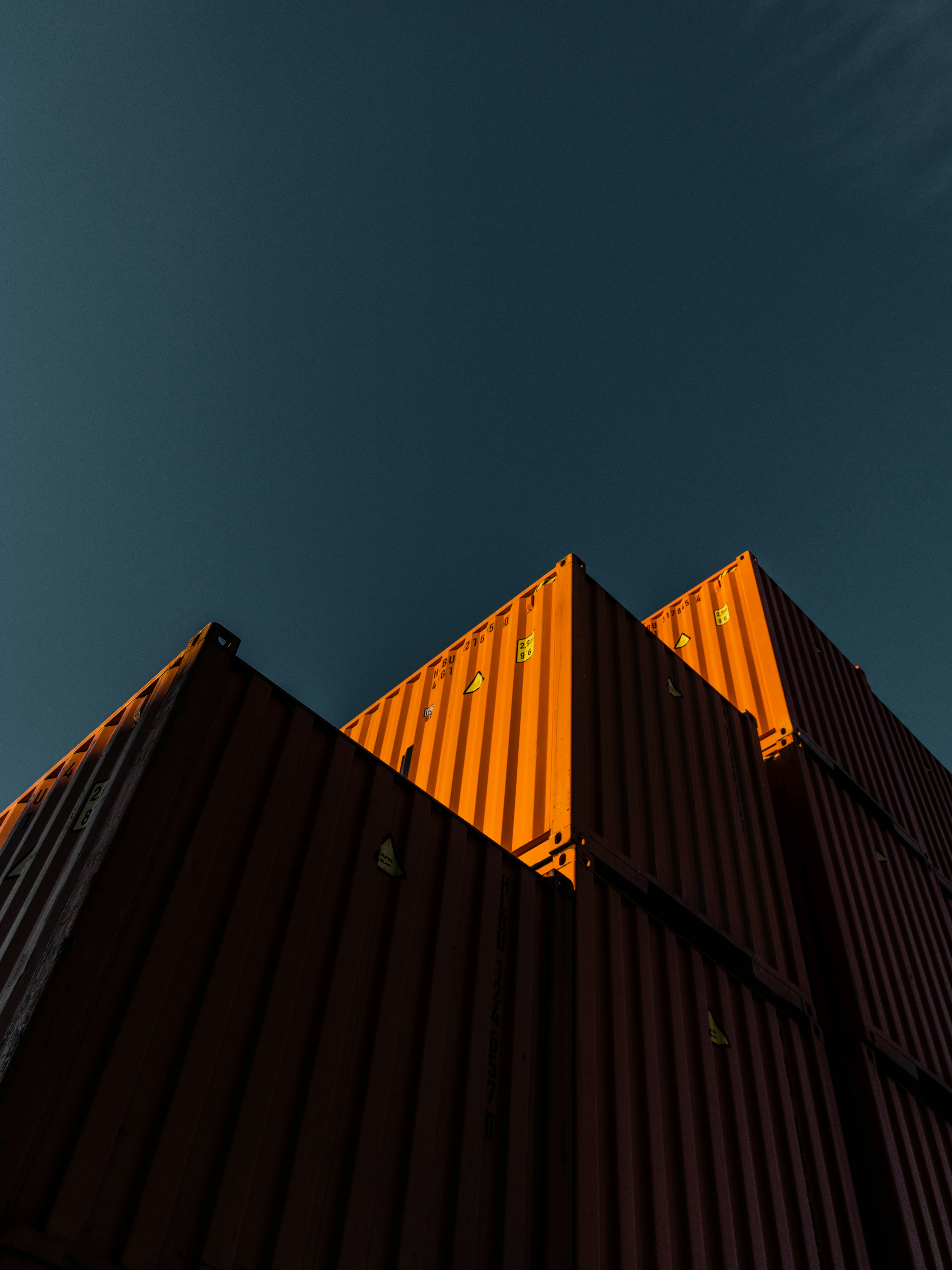 Stack of orange shipping containers illuminated by sunlight against a dark blue sky.