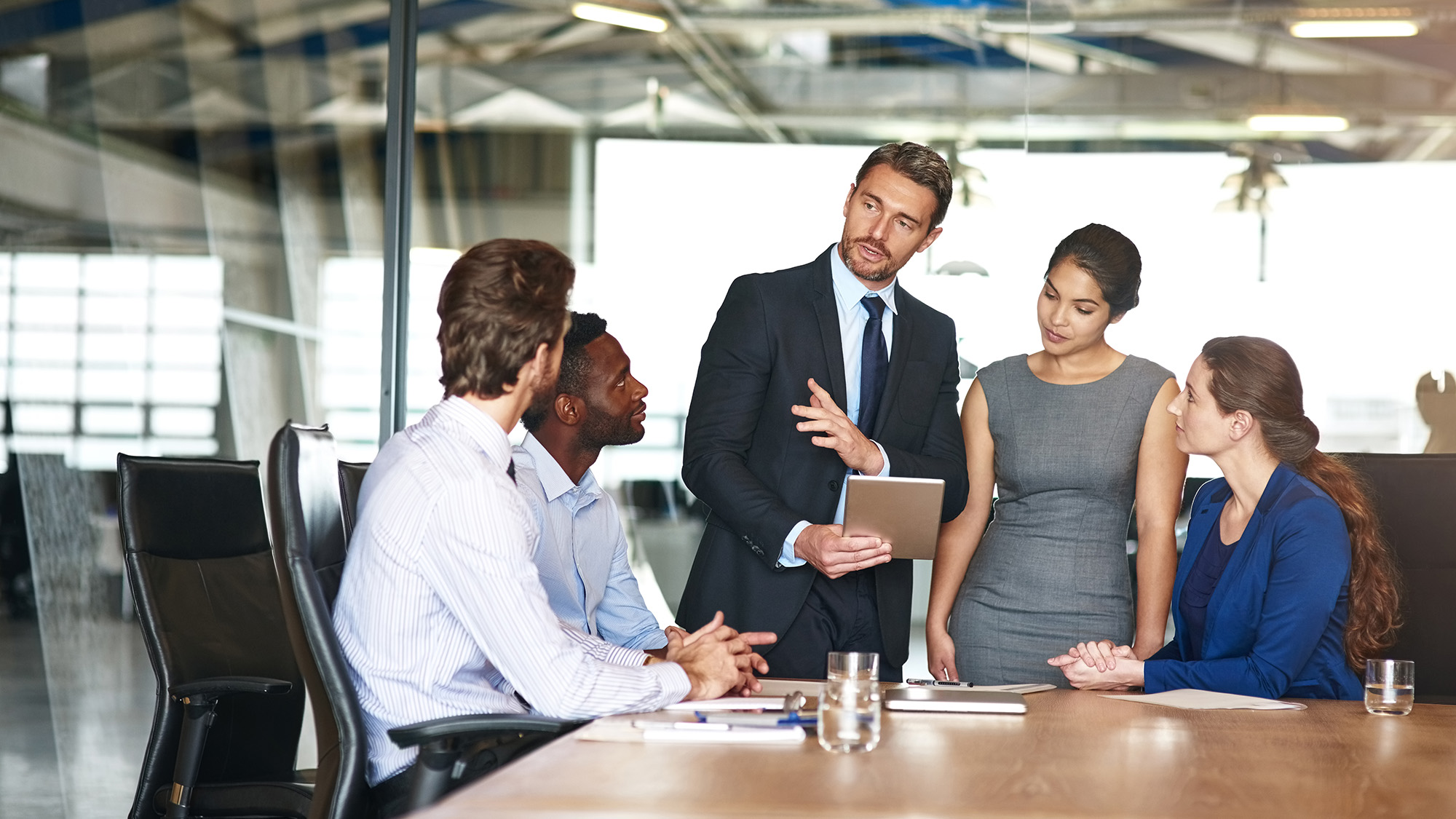 Man presenting tablet to colleagues in a glass-walled conference room