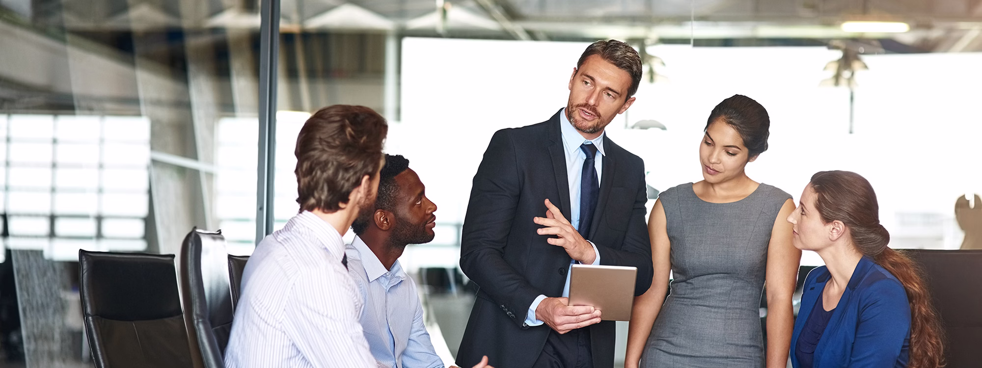 Man presenting tablet to colleagues in a glass-walled conference room