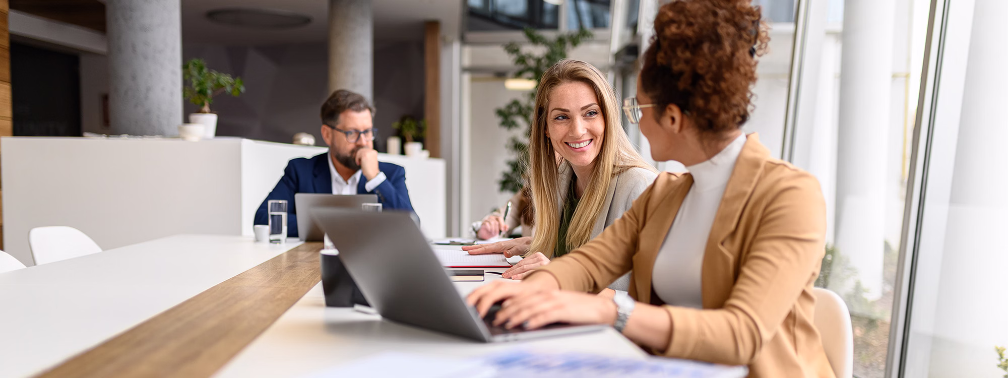 Two women converse while using laptop at long meeting table in office