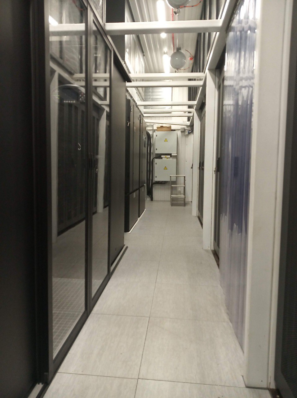 Interior corridor of energy-efficient modular data center with server racks behind glass doors, raised floor tiles, and power distribution panels