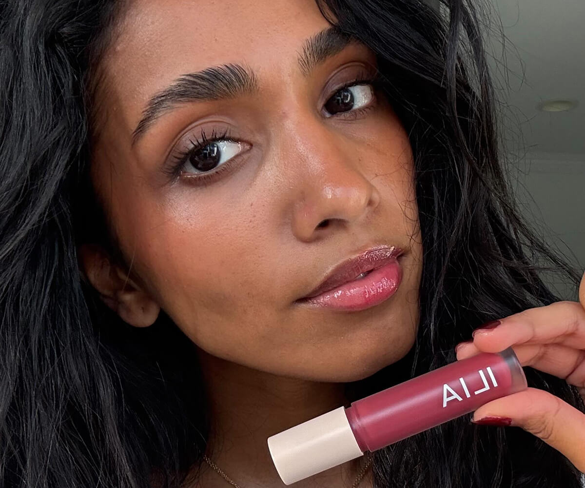 Close-up of a woman with dark hair holding a pink ILIA lip gloss near her glossy lips.