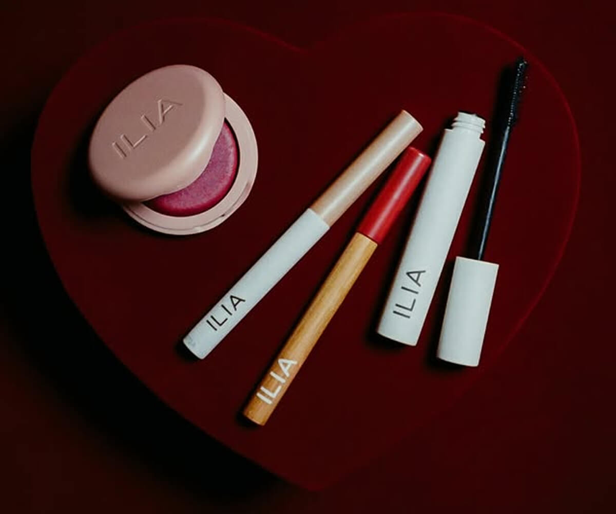 ILIA cosmetic products including blush compact, lipsticks, and mascara arranged on a red heart-shaped surface.