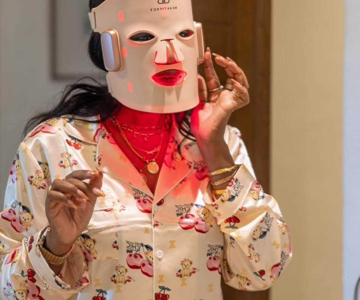 Person wearing a light therapy LED facial mask with red lights and a patterned silk pajama top.